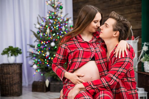 A husband holds his pregnant wife on his lap. The couple looks straight into each other's eyes. The spouses are wearing identical red checkered pajamas. In the background stands a Christmas tree and a potted plant.