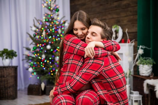 A couple dressed in identical red plaid pajamas cuddle affectionately as the woman sits on the man's lap. In the background is a smudged Christmas tree with Christmas lights.