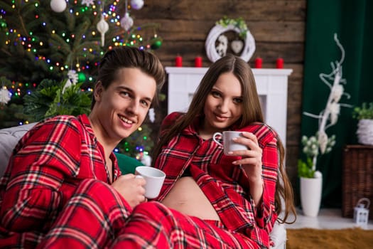 A woman with an exposed pregnancy belly and her partner sit on a gray couch against a backdrop of a Christmas tree and holiday decorations. The couple looks ahead toward the lens. The individuals are dressed in identical plaid pajamas.