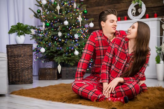 A couple sits on a fluffy brown carpet and makes funny faces. The man sits at the back and the woman in front of him. In the background is a dressed Christmas tree and other holiday decorations.