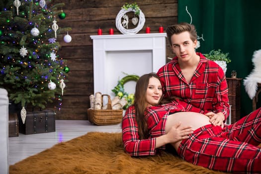 A pregnant woman lies on a brown fluffy carpet. She is resting her back on the knees of her husband kneeling next to her. There is a Christmas tree and other holiday decorations in the room.