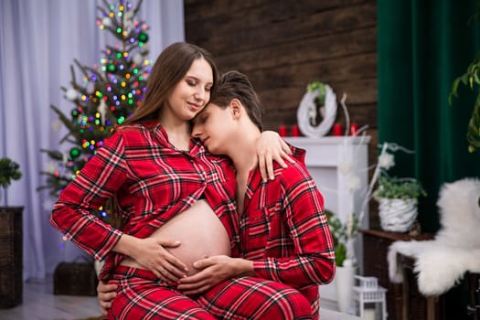 A couple in love with their eyes closed cuddle affectionately as the woman sits on the man's lap. The man snuggles his face into his partner's shoulder. Both hold their hands on the pregnant belly.