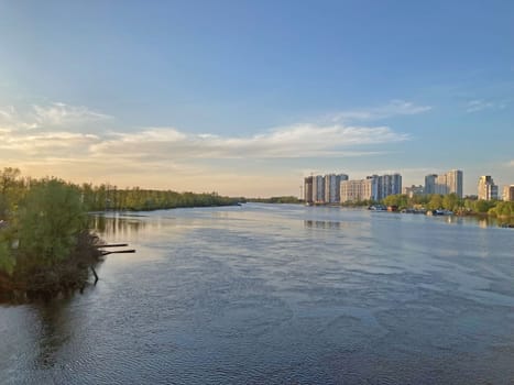 Beautiful panoramic view of the city of Kyiv Dneper River at sunset on dark blue water with glowing reflected rays of the sun, copy-space. View of the deep river with the buildings in the background.