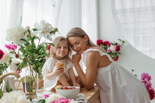 A little blonde girl with her mom on a kitchen countertop decorated with peonies. The concept of the relationship between mother and daughter. Spring atmosphere.