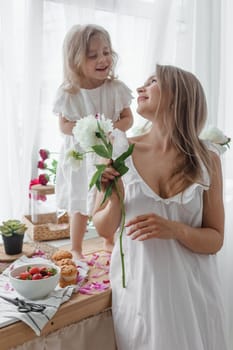 A little blonde girl with her mom on a kitchen countertop decorated with peonies. The concept of the relationship between mother and daughter. Spring atmosphere.