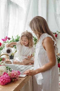 A little blonde girl with her mom on a kitchen countertop decorated with peonies. The concept of the relationship between mother and daughter. Spring atmosphere.