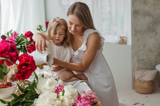 A little blonde girl with her mom on a kitchen countertop decorated with peonies. The concept of the relationship between mother and daughter. Spring atmosphere.