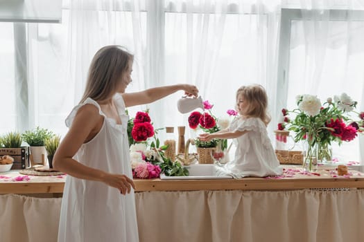 A little blonde girl with her mom on a kitchen countertop decorated with peonies. The concept of the relationship between mother and daughter. Spring atmosphere.