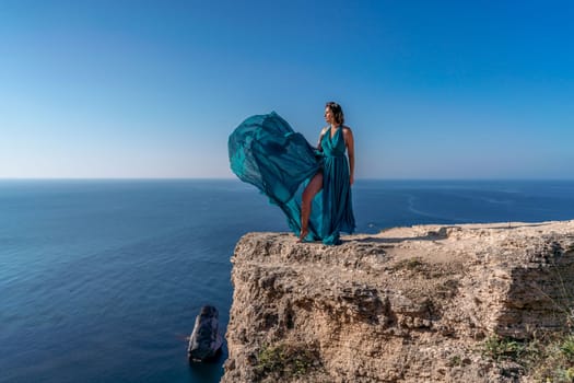 A girl with loose hair in a long mint dress descends the stairs between the yellow rocks overlooking the sea. A rock can be seen in the sea. Sunny path on the sea from the rising sun.