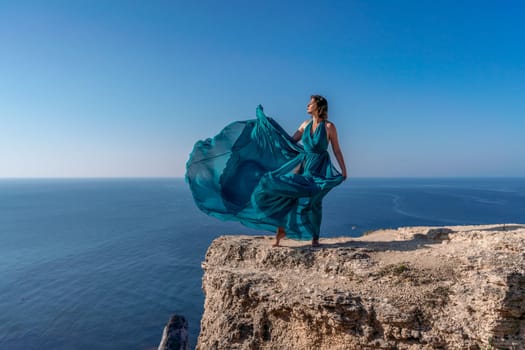 A girl with loose hair in a long mint dress descends the stairs between the yellow rocks overlooking the sea. A rock can be seen in the sea. Sunny path on the sea from the rising sun.