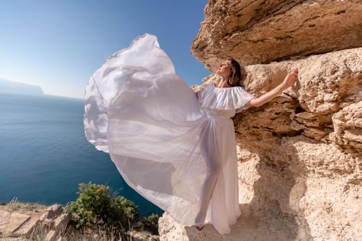 A beautiful young woman in a white light dress with long legs stands on the edge of a cliff above the sea waving a white long dress, against the background of the blue sky and the sea