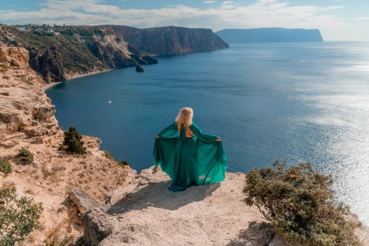 A beautiful young woman in a mint light dress with long legs stands on the edge of a cliff above the sea waving long dress, against the background of the blue sky and the sea