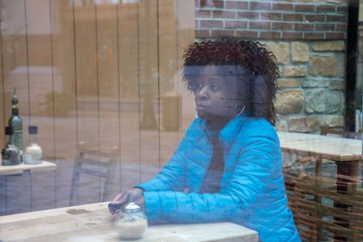 An African American woman in a blue jacket sits behind a window glass in a cafe waiting for a waiter, selective soft focus of the photo due to dirty glass. High quality photo