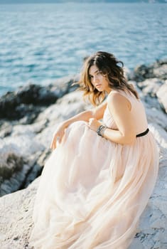 Bride in a beige dress with tousled hair sits on the rocks by the sea. High quality photo