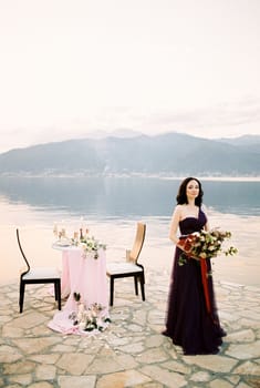 Woman with a bouquet of flowers stands near the laid festive table on the pier. High quality photo