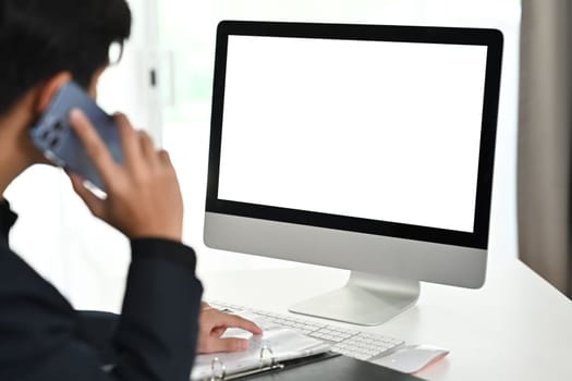Cropped image of young male investor having phone conversation and looking at computer monitor at office desk.