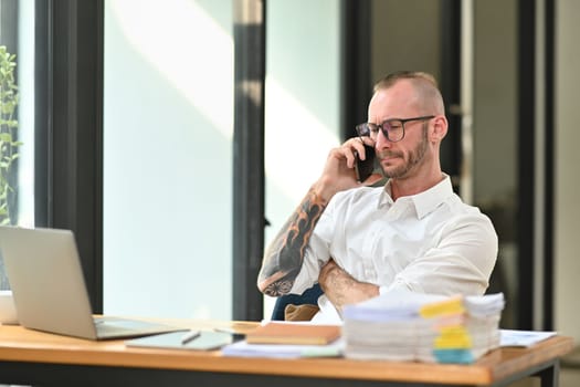 Confident handsome businessman in a white shirt with tattooed talking mobile phone and using laptop at his workplace.