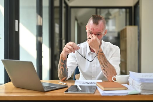 Tired caucasian man office worker holding glasses and rubbing nose suffering from chronic dry eye syndrome bridge.