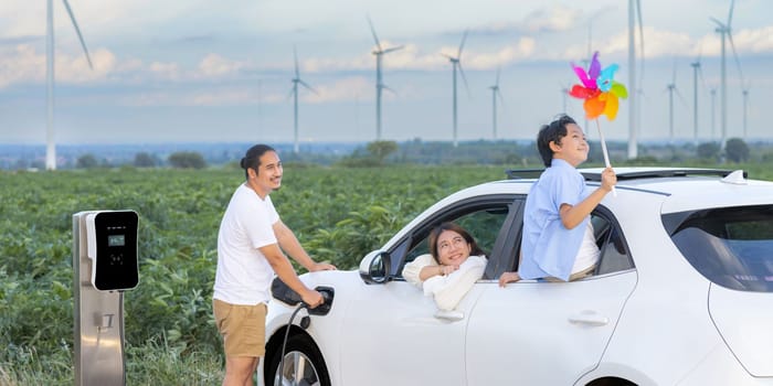 Concept of progressive happy family enjoying their time at wind farm with electric vehicle. Electric vehicle driven by clean renewable energy from wind turbine generator for charging station.