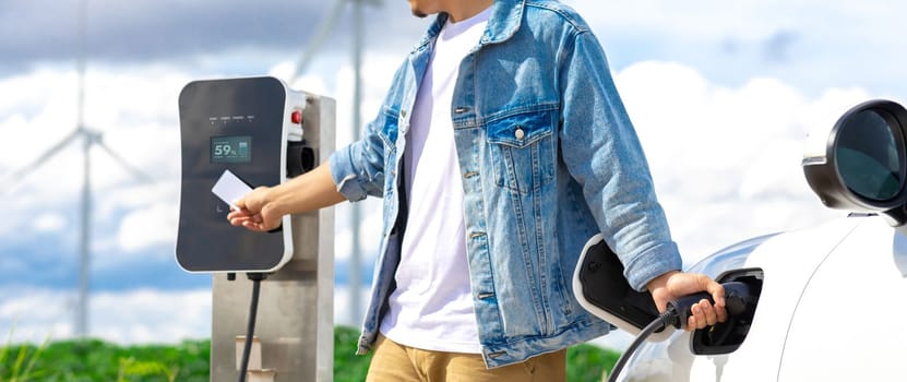 Progressive man with his electric car, EV car recharging energy from charging station on green field with wind turbine as concept of future sustainable energy. Electric vehicle with energy generator.