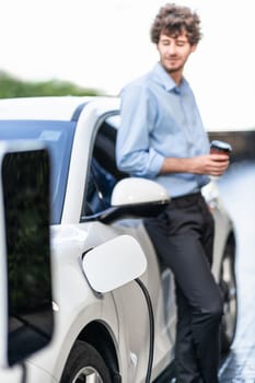 Progressive eco-friendly concept of focus parking EV car at public electric-powered charging station in city with blur background of businessman leaning on recharging-electric vehicle with coffee.