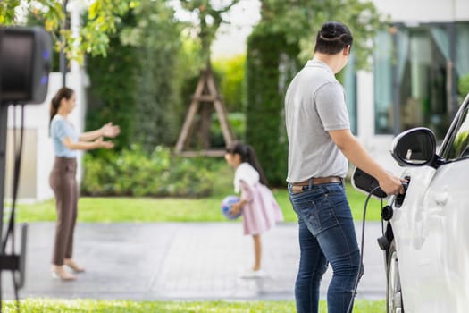 Focus image of progressive man charging electric car from home charging station with blur mother and daughter playing together in the background.
