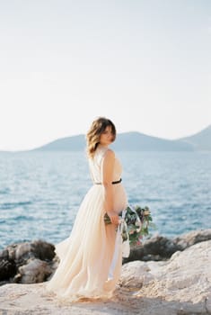 Bride with a bouquet stands sideways on a rocky seashore. High quality photo