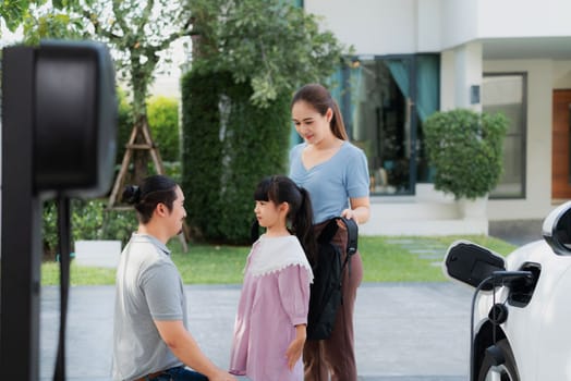 Progressive young parents and daughter with electric vehicle and home charging station. Green and clean energy from electric vehicles for healthy environment. Eco power from renewable source at home.