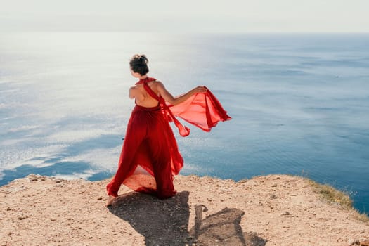 Side view a Young beautiful sensual woman in a red long dress posing on a rock high above the sea during sunrise. Girl on the nature on blue sky background. Fashion photo.