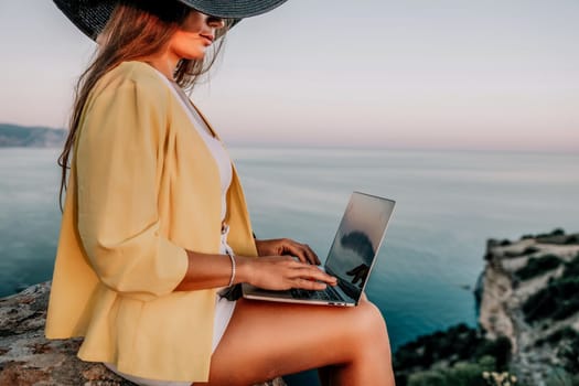 Successful business woman in yellow hat working on laptop by the sea. Pretty lady typing on computer at summer day outdoors. Freelance, travel and holidays concept.