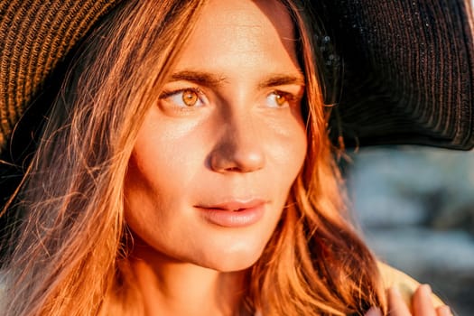 Portrait of happy young woman wearing summer black hat with large brim at beach on sunset. Closeup face of attractive girl with black straw hat. Happy young woman smiling and looking at camera at sea