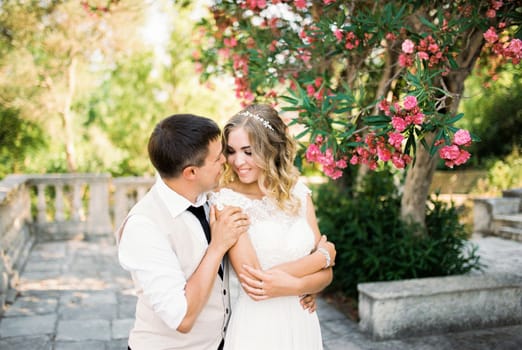 Groom hugs bride from behind by the shoulders under a blooming oleander. High quality photo