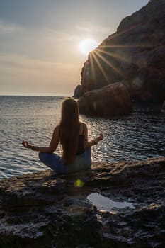 Woman tourist enjoying the sunset over the sea mountain landscape. Sits outdoors on a rock above the sea. She is wearing jeans and a blue hoodie. Healthy lifestyle, harmony and meditation.