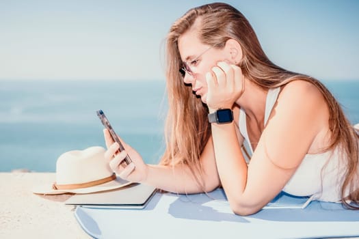 Successful business woman in yellow hat working on laptop by the sea. Pretty lady typing on computer at summer day outdoors. Freelance, travel and holidays concept.