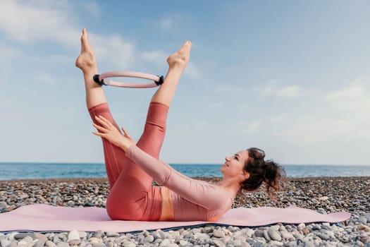 Middle aged well looking woman with black hair doing Pilates with the ring on the yoga mat near the sea on the pebble beach. Female fitness yoga concept. Healthy lifestyle, harmony and meditation.