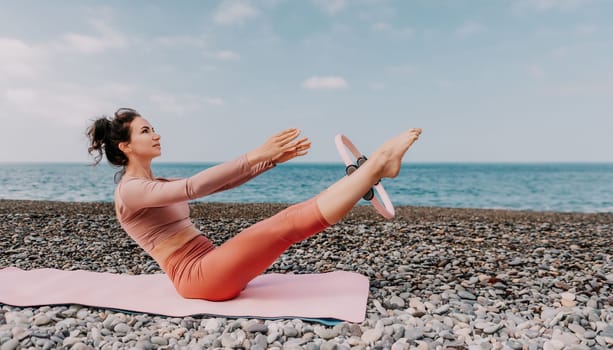 Middle aged well looking woman with black hair doing Pilates with the ring on the yoga mat near the sea on the pebble beach. Female fitness yoga concept. Healthy lifestyle, harmony and meditation.
