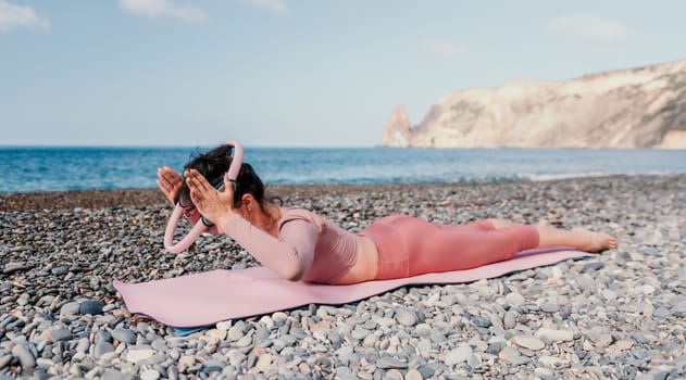 Middle aged well looking woman with black hair doing Pilates with the ring on the yoga mat near the sea on the pebble beach. Female fitness yoga concept. Healthy lifestyle, harmony and meditation.