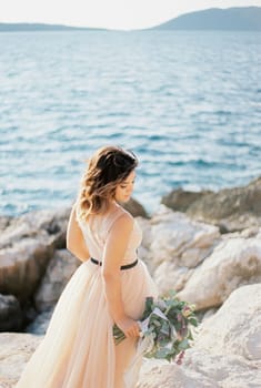 Bride with a bouquet stands sideways with her head bowed on a rocky seashore. High quality photo