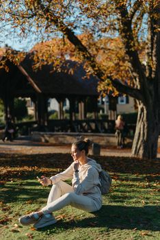 Young fashionable teenage girl with smartphone in park in autumn sitting at smiling. Trendy young woman in fall in park texting. Retouched, vibrant colors. Beautiful blonde teenage girl wearing casual modern autumn outfit sitting in park in autumn. Retouched, vibrant colors, brownish tones.