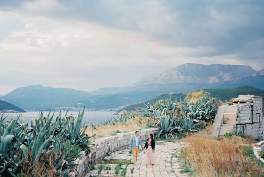 Bride and groom walk, holding hands, along the stone road past the ruins of the fortress. High quality photo