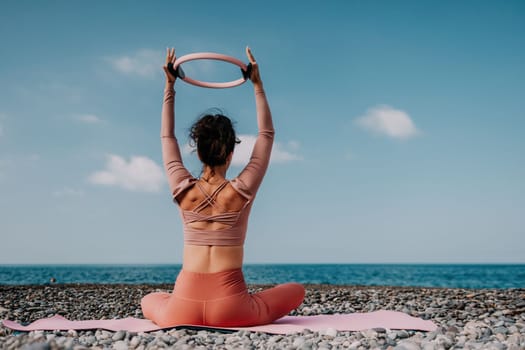 Middle aged well looking woman with black hair doing Pilates with the ring on the yoga mat near the sea on the pebble beach. Female fitness yoga concept. Healthy lifestyle, harmony and meditation.