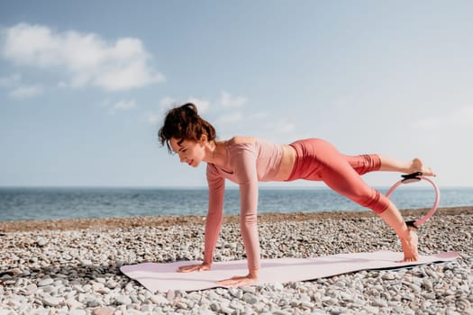 Middle aged well looking woman with black hair doing Pilates with the ring on the yoga mat near the sea on the pebble beach. Female fitness yoga concept. Healthy lifestyle, harmony and meditation.