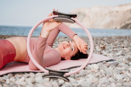 Middle aged well looking woman with black hair doing Pilates with the ring on the yoga mat near the sea on the pebble beach. Female fitness yoga concept. Healthy lifestyle, harmony and meditation.