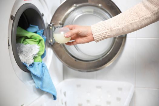 Female hands pouring liquid laundry detergent into measure cup. Washing machine and clothes in white modern room close up
