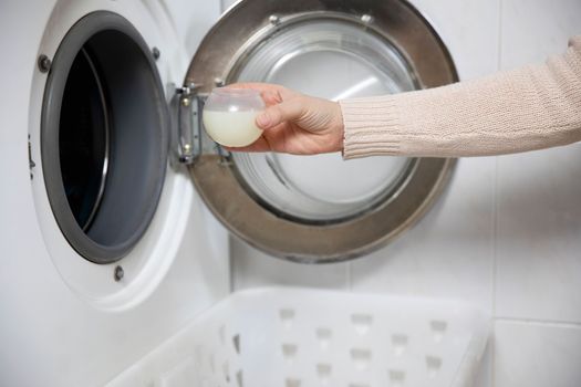 Female hands pouring liquid laundry detergent into measure cup. Washing machine and clothes in white modern room close up