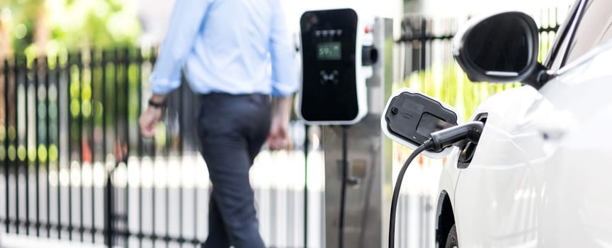 Focus closeup electric vehicle recharge battery at public charging station in the city area with blur businessman in background. EV car attached with electric charger for eco-friendly idea.