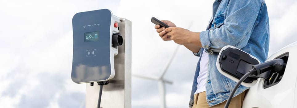 Progressive man with his electric car, EV car recharging energy from charging station on green field with wind turbine as concept of future sustainable energy. Electric vehicle with energy generator.