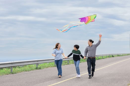 Progressive happy family vacation and carefree day concept. Young parents mother father and son run along and flying kite together road with enjoy natural scenic on scenery and clear sky background.