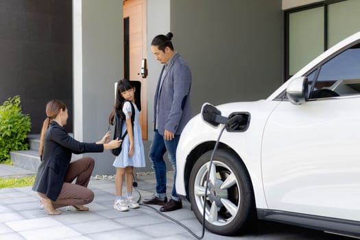 Progressive young parents and daughter with electric vehicle and home charging station. Green and clean energy from electric vehicles for healthy environment. Eco power from renewable source at home.