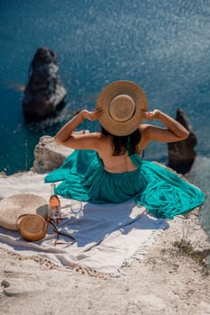 A girl in a hat sits with her back to the viewer on a picnic blanket in a mint dress and holds a hat with her hands. Champagne, two glasses, Summit on the mountain against the background of the sea and rocks in the sea